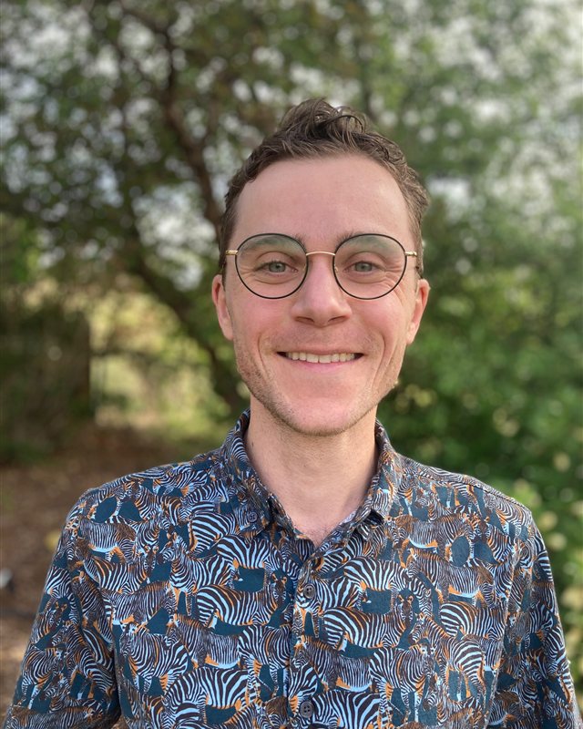 Sam Haraway, smiling, outdoors in front of green trees